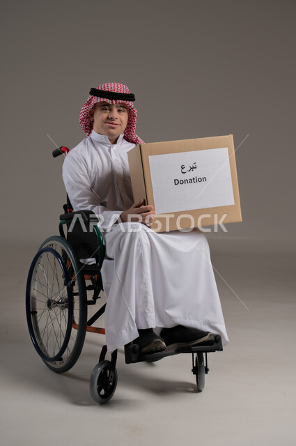 مفهوم مساعدة المحتاجين, ذوي الإحتياجات الخاصة والهمم, بورتريه لشاب عربي خليجي سعودي يرتدي الثوب والشماغ يجلس على كرسي متحرك ويمسك بيديه صندوق تبرع, خلفية رمادي