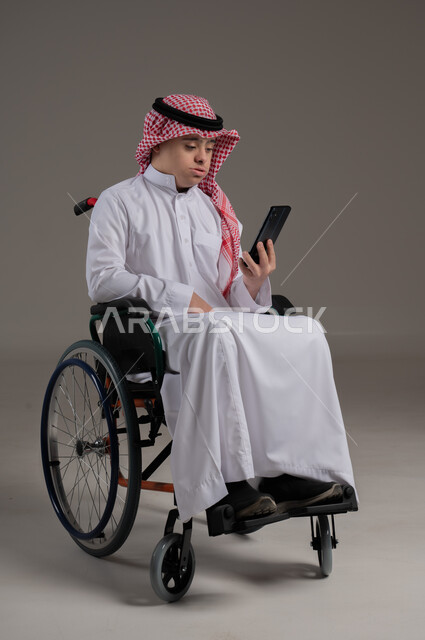 مفهوم التحدث مع الاصدقاء والاهل بواسطة برامج التواصل الاجتماعي, ذوي الإحتياجات الخاصة والهمم, بورتريه لشاب عربي خليجي سعودي يرتدي الثوب والشماغ يجلس على كرسي متحرك ويمسك بيده هاتف محمول, خلفية رمادي