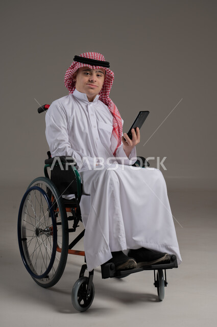 مفهوم التحدث مع الاصدقاء والاهل بواسطة برامج التواصل الاجتماعي, ذوي الإحتياجات الخاصة والهمم, بورتريه لشاب عربي خليجي سعودي يرتدي الثوب والشماغ يجلس على كرسي متحرك ويمسك بيده هاتف محمول, خلفية رمادي