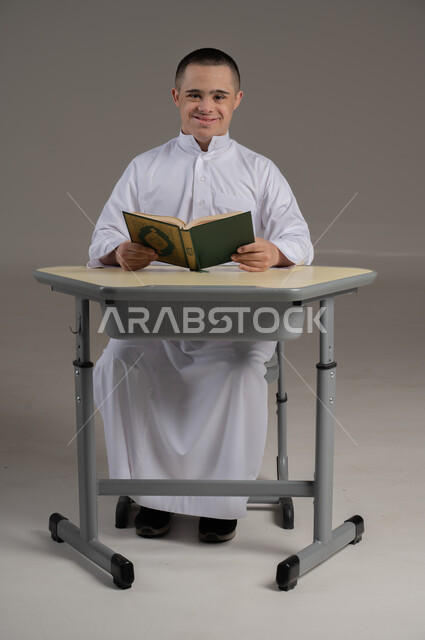 تنمية قدرات الأشخاص ذوي الاحتياجات الخاصة، تلاوة المصحف الشريف، بورتريه لشاب عربي خليجي عماني مصاب بمتلازمة داون يرتدي الدشداشة يمسك بيده القرآن، قراءة كتاب الله وحفظه وتجويده، مفهوم الاجتهاد في العبادة، ايماءات السرور والاطمئنان، خلفية رمادية