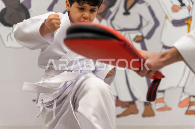 فتى عربي خليجي سعودي يقوم بالتدرب على الحركات القتالية  بمساعدة مدرب خليجي سعودي خاص برياضة الكارتيه اليابانية باستخدام أداة تدريبية لتطوير مهارتي الركل واللكم ​، التدرب على فنون الدفاع عن النفس ، رياضة دفاعية تتميز بالأخلاق العالية للاعبين