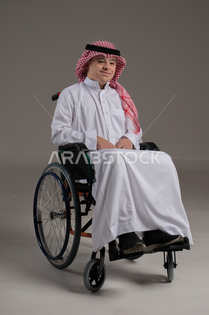 تعابير السعادة والفرح, ذوي الإحتياجات الخاصة والهمم, بورتريه لشاب عربي خليجي سعودي يرتدي الثوب والشماغ يجلس على كرسي متحرك, خلفية رمادي