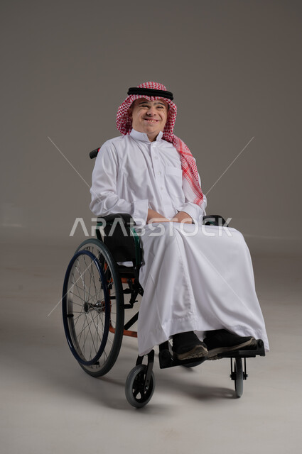 تعابير السعادة والفرح, ذوي الإحتياجات الخاصة والهمم, بورتريه لشاب عربي خليجي سعودي يرتدي الثوب والشماغ يجلس على كرسي متحرك, خلفية رمادي