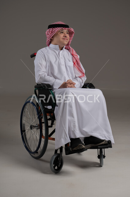 Expressions of happiness and joy, people with special needs and concerns, portrait of a Saudi Gulf Arab young man wearing a thobe and shemagh sitting in a wheelchair, gray background