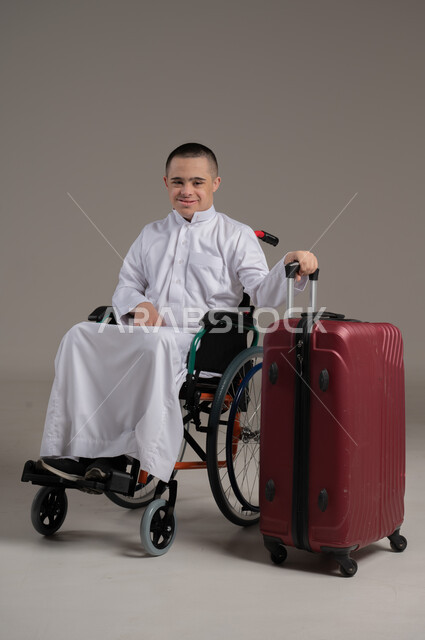 مفهوم الاسترخاء والاستراحة, بورتريه لولد عربي خليجي عماني مقعد مصاب بمتلازمة داون يرتدي الدشداشه يجلس بجانب حقيبة السفر, النظر الى الكاميرا بايماءات السرور، قضاء وقت الإجازة في السياحة والسفر، صورة الجسم بالطول الكامل, خلفية رمادية