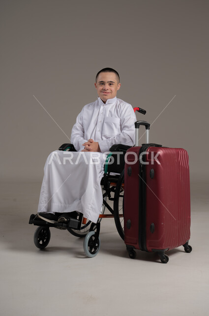 مفهوم الاسترخاء والاستراحة, بورتريه لولد عربي خليجي عماني مقعد مصاب بمتلازمة داون يرتدي الدشداشه يجلس بجانب حقيبة السفر, النظر الى الكاميرا بايماءات السرور، قضاء وقت الإجازة في السياحة والسفر، صورة الجسم بالطول الكامل, خلفية رمادية
