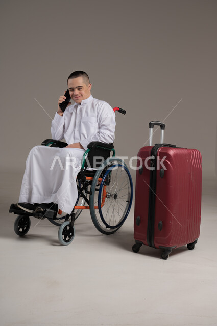 التواصل مع الاصدقاء عبر الهاتف المحمول، قضاء وقت الإجازة في السياحة والسفر، مفهوم الاسترخاء والاستراحة, بورتريه لولد عربي خليجي عماني مقعد مصاب بمتلازمة داون يرتدي الدشداشه يجلس بجانب حقيبة السفر يقوم باجراء مكالمة هاتفية عبر الجوال، صورة لكامل الجسم, خلفية رمادية