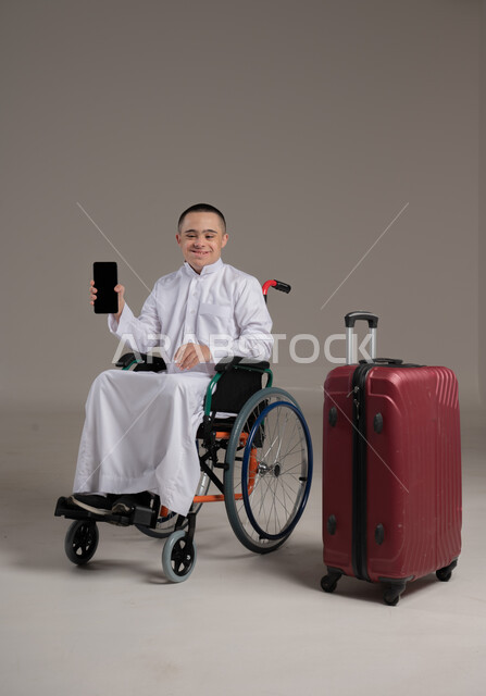 عرض شاشة سوداء فارغة، استخدام الاجهزة الحديثة، قضاء وقت الإجازة في السياحة والسفر، مفهوم الاسترخاء والاستراحة, بورتريه لولد عربي خليجي عماني مقعد مصاب بمتلازمة داون يرتدي الدشداشه يجلس بجانب حقيبة السفر يحمل بيده الهاتف المحمول، صورة لكامل الجسم, خلفية رمادية