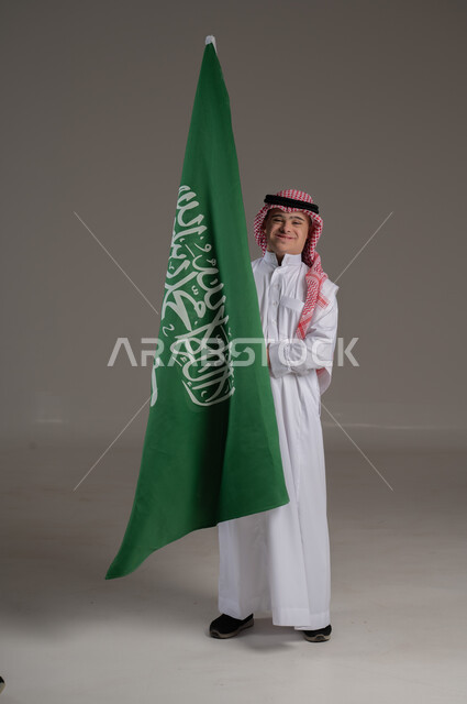 مفهوم حب الوطن والانتماء اليه, ذوي الاحتياجات الخاصة والهمم, بورتريه لشاب عربي خليجي سعودي يرتدي الثوب والشماغ يمسك علم المملكة العربية السعودية بايماءات الفخر والاعتزاز, صورة الجسم بالطول الكامل, خلفية رمادي