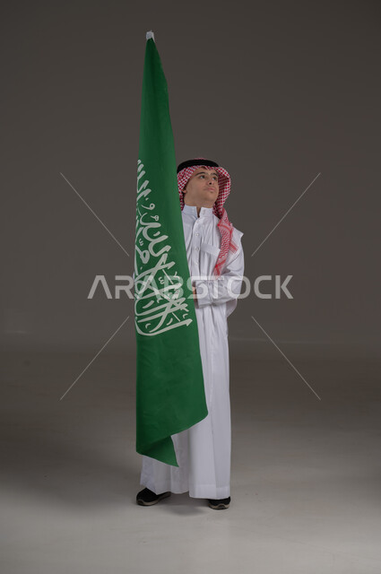 مفهوم حب الوطن والانتماء اليه, ذوي الاحتياجات الخاصة والهمم, بورتريه لشاب عربي خليجي سعودي يرتدي الثوب والشماغ يمسك علم المملكة العربية السعودية بايماءات الفخر والاعتزاز, صورة الجسم بالطول الكامل, خلفية رمادي