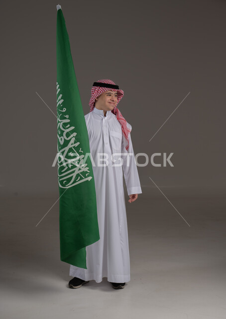 مفهوم حب الوطن والانتماء اليه, ذوي الاحتياجات الخاصة والهمم, بورتريه لشاب عربي خليجي سعودي يرتدي الثوب والشماغ يمسك علم المملكة العربية السعودية بايماءات الفخر والاعتزاز, صورة الجسم بالطول الكامل, خلفية رمادي