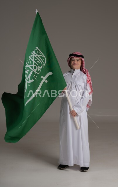 مفهوم حب الوطن والانتماء اليه, ذوي الاحتياجات الخاصة والهمم, بورتريه لشاب عربي خليجي سعودي يرتدي الثوب والشماغ يمسك علم المملكة العربية السعودية بايماءات الفخر والاعتزاز, صورة الجسم بالطول الكامل, خلفية رمادي