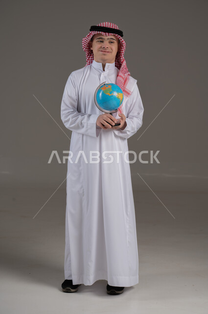 النظر الى الكاميرا بتعابير السعادة والسرور, ذوي الاحتياجات الخاصة والهمم, بورتريه لشاب عربي خليجي سعودي يرتدي الثوب والشماغ يمسك بيديه كرة ارضية, صورة الجسم بالطول الكامل, خلفية رمادي
