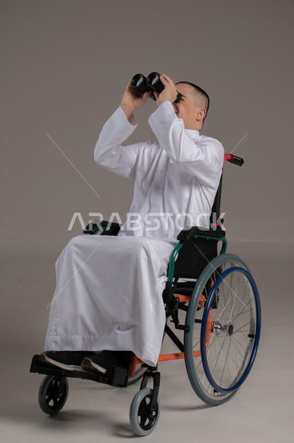 التخطيط المستقبلي للأهداف والطموحات، حب الاستكشاف والمغامرة، رؤية مستقبلية عند أصحاب ذوي الهمم، بورتريه لولد عربي خليجي عماني مقعد مصاب بمتلازمة داون يرتدي الدشداشة يجلس على كرسي متحرك و يحمل بيده المنظار ثنائي العينين، خلفية رمادية