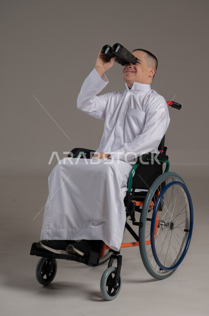 رؤية مستقبلية عند أصحاب ذوي الهمم، التخطيط المستقبلي للأهداف والطموحات، حب الاستكشاف والمغامرة، بورتريه لولد عربي خليجي عماني مقعد مصاب بمتلازمة داون يرتدي الدشداشة يجلس على كرسي متحرك و يحمل بيده المنظار ثنائي العينين، خلفية رمادية