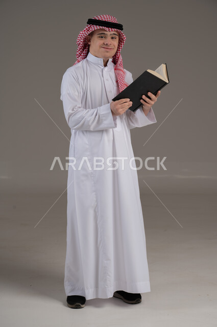 مفهوم القراءة والتعلم والمطالعة, ذوي الاحتياجات الخاصة والهمم, بورتريه لشاب عربي خليجي سعودي يرتدي الثوب والشماغ يمسك بيديه كتاب, صورة الجسم بالطول الكامل, خلفية رمادي