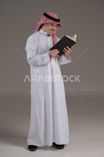مفهوم القراءة والتعلم والمطالعة, ذوي الاحتياجات الخاصة والهمم, بورتريه لشاب عربي خليجي سعودي يرتدي الثوب والشماغ يمسك بيديه كتاب, صورة الجسم بالطول الكامل, خلفية رمادي
