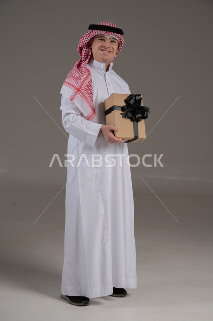 اعياد الميلاد والمناسبات, مفهوم تقديم الهداية, ذوي الاحتياجات الخاصة والهمم, بورتريه لشاب عربي خليجي سعودي يرتدي الثوب والشماغ يمسك هدية, صورة الجسم بالطول الكامل, خلفية رمادي