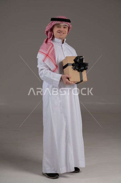 اعياد الميلاد والمناسبات, مفهوم تقديم الهداية, ذوي الاحتياجات الخاصة والهمم, بورتريه لشاب عربي خليجي سعودي يرتدي الثوب والشماغ يمسك هدية, صورة الجسم بالطول الكامل, خلفية رمادي