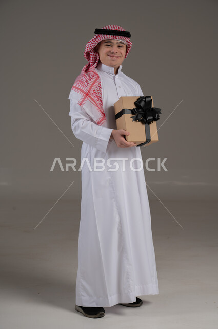اعياد الميلاد والمناسبات, مفهوم تقديم الهداية, ذوي الاحتياجات الخاصة والهمم, بورتريه لشاب عربي خليجي سعودي يرتدي الثوب والشماغ يمسك هدية, صورة الجسم بالطول الكامل, خلفية رمادي
