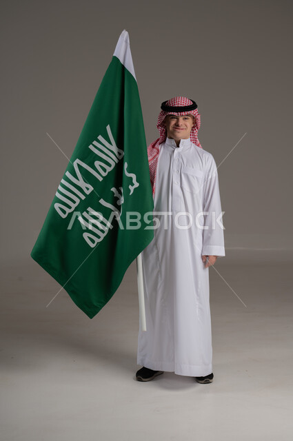 مفهوم حب الوطن والانتماء اليه, ذوي الاحتياجات الخاصة والهمم, بورتريه لشاب عربي خليجي سعودي يرتدي الثوب والشماغ يمسك بيده علم المملكة العربية السعودية بتعابير الفخر والاعتزاز, صورة الجسم بالطول الكامل, خلفية رمادي