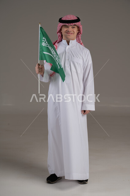 مفهوم حب الوطن والانتماء اليه, ذوي الاحتياجات الخاصة والهمم, بورتريه لشاب عربي خليجي سعودي يرتدي الثوب والشماغ يمسك بيده علم المملكة العربية السعودية بتعابير الفخر والاعتزاز, صورة الجسم بالطول الكامل, خلفية رمادي
