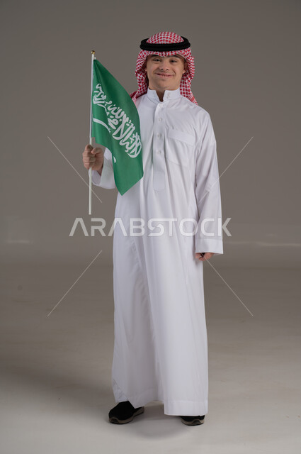 مفهوم حب الوطن والانتماء اليه, ذوي الاحتياجات الخاصة والهمم, بورتريه لشاب عربي خليجي سعودي يرتدي الثوب والشماغ يمسك بيده علم المملكة العربية السعودية بتعابير الفخر والاعتزاز, صورة الجسم بالطول الكامل, خلفية رمادي