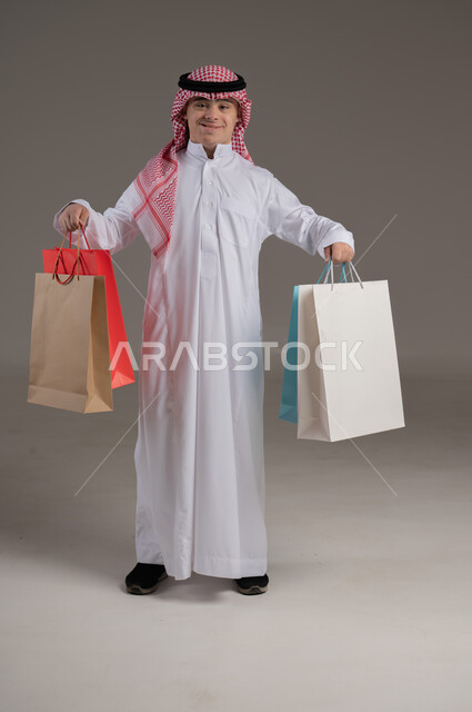 مفهوم التسوق والتبضع, ذوي الاحتياجات الخاصة والهمم, بورتريه لشاب عربي خليجي سعودي يرتدي الثوب والشماغ يمسك بيديه مجموعة من الاكياس, صورة الجسم بالطول الكامل, خلفية رمادي