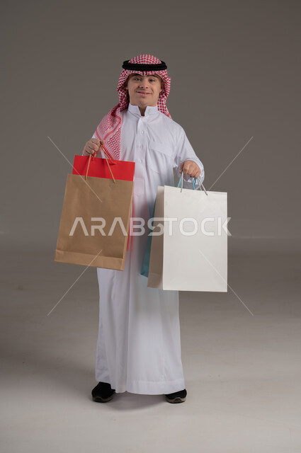 مفهوم التسوق والتبضع, ذوي الاحتياجات الخاصة والهمم, بورتريه لشاب عربي خليجي سعودي يرتدي الثوب والشماغ يمسك بيديه مجموعة من الاكياس, صورة الجسم بالطول الكامل, خلفية رمادي