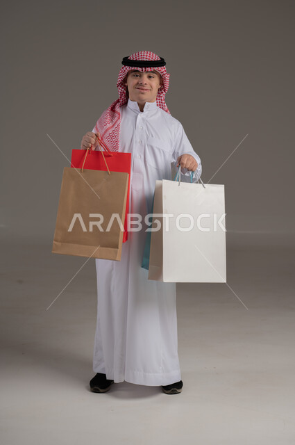 مفهوم التسوق والتبضع, ذوي الاحتياجات الخاصة والهمم, بورتريه لشاب عربي خليجي سعودي يرتدي الثوب والشماغ يمسك بيديه مجموعة من الاكياس, صورة الجسم بالطول الكامل, خلفية رمادي
