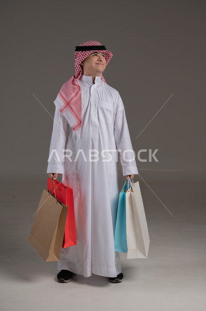 مفهوم التسوق والتبضع, ذوي الاحتياجات الخاصة والهمم, بورتريه لشاب عربي خليجي سعودي يرتدي الثوب والشماغ يمسك بيديه مجموعة من الاكياس, صورة الجسم بالطول الكامل, خلفية رمادي