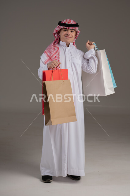 مفهوم التسوق والتبضع, ذوي الاحتياجات الخاصة والهمم, بورتريه لشاب عربي خليجي سعودي يرتدي الثوب والشماغ يمسك بيديه مجموعة من الاكياس, صورة الجسم بالطول الكامل, خلفية رمادي