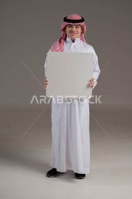 النظر الى الكاميرا بتعابير السعادة والسرور, ذوي الاحتياجات الخاصة والهمم, بورتريه لشاب عربي خليجي سعودي يرتدي الثوب والشماغ يمسك بيديه لوح ابيض فارغ, صورة الجسم بالطول الكامل, خلفية رمادي