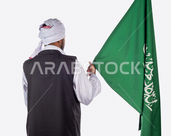بورتريه بزاوية من الخلف لرجل عربي سوداني، يحمل بيده علم المملكة العربية السعودية، ذكرى اليوم الوطني السعودي، الاحتفال بالعيد الوطني السعودي، خلفية بيضاء لرجل سوداني مسلم يرتدي الزي السوداني مع العمه الملفحة