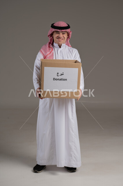 النظر الى الكاميرا بتعابير السعادة والسرور, مفهوم مساعدة المحتاجين, ذوي الاحتياجات الخاصة والهمم, بورتريه لشاب عربي خليجي سعودي يرتدي الثوب والشماغ يمسك بيديه صندوق تبرع, صورة الجسم بالطول الكامل, خلفية رمادي