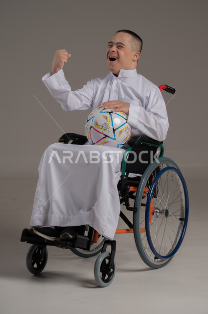 تنمية الهوايات الرياضية لذوي الإعاقة, بورتريه لشاب عربي خليجي عماني مبتسم مقعد مصاب بمتلازمة داون يجلس على كرسي متحرك يحمل بيده كرة القدم، ايماءات الفوز والنجاح، تعابير الفرح والسرور، نشاطات ترفيهية لذوي الهمم، خلفية رمادية