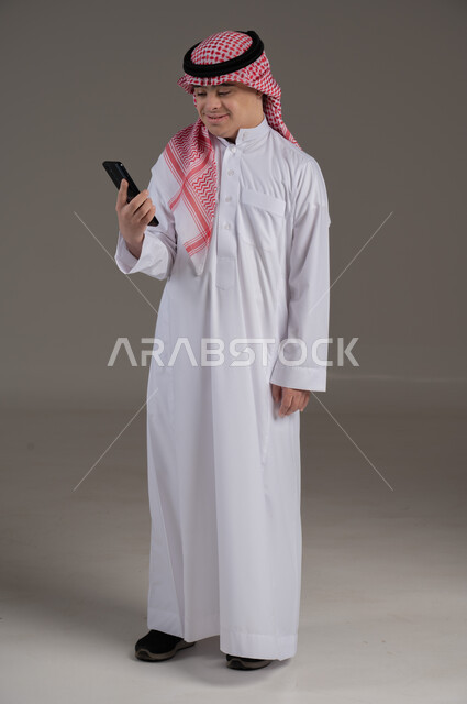 مفهوم التواصل مع العائلة والاصدقاء, ذوي الاحتياجات الخاصة والهمم, بورتريه لشاب عربي خليجي سعودي يرتدي الثوب والشماغ يمسك بيده هاتف محمول, صورة الجسم بالطول الكامل, خلفية رمادي