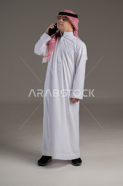 مفهوم التواصل مع العائلة والاصدقاء, ذوي الاحتياجات الخاصة والهمم, بورتريه لشاب عربي خليجي سعودي يرتدي الثوب والشماغ يمسك بيده هاتف محمول, صورة الجسم بالطول الكامل, خلفية رمادي