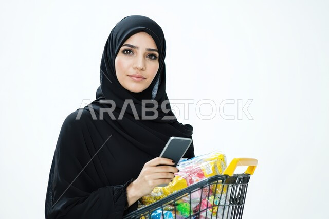 شراء الاحتياجات و المستلزمات المنزلية للمستهلك، صورة مقربة لامرأة عربية سعودية خليجية محجبة مبتسمة ترتدي العباءة السوداء تنظر الى الكاميرا بإيماءات الثقة تحمل بيدها سلة التسوق تقوم بالتبضع في احدى أسواق المملكة العربية السعودية، السلع والمواد الغذائية المختلفة، خلفية بيضاء