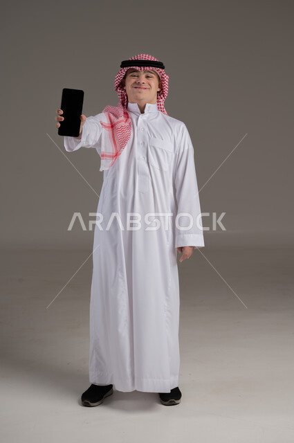 النظر الى الكاميرا بتعابير السعادة والسرور, ذوي الاحتياجات الخاصة والهمم, بورتريه لشاب عربي خليجي سعودي يرتدي الثوب والشماغ يمسك بيده هاتف محمول بشاشة سوداء, صورة الجسم بالطول الكامل, خلفية رمادي