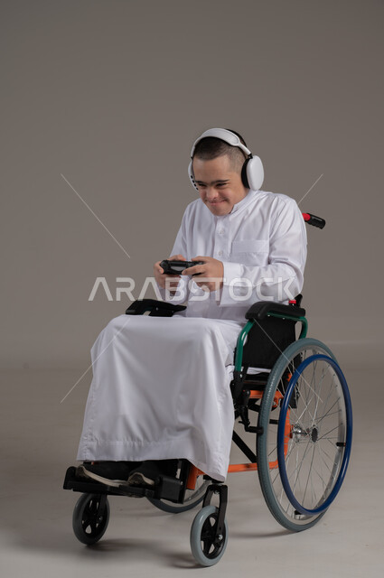 Recreational activities for people with disabilities, gestures of joy and happiness, playing PlayStation games, fun and entertainment with video games, portrait of a disabled Gulf Arab Omani boy with Down syndrome wearing a dishdasha and headphones holding a joystick in his hand, full body image, gray background