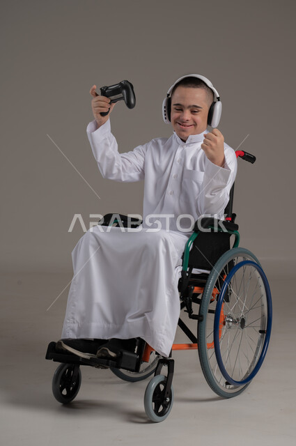 لعب ألعاب البلايستيشن، المتعة والتسلية بألعاب الفيديو، بورتريه لولد عربي خليجي عماني مقعد مصاب بمتلازمة داون يمسك بيده عصا التحكم يرفع يده بايماءات الفوز، الاهتمام بقدرات الأشخاص ذوي الاحتياجات الخاصة، نشاطات ترفيهية لذوي الهمم، صورة لكامل الجسم، خلفية رمادية