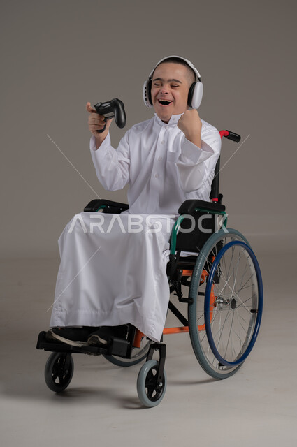 المتعة والتسلية بألعاب الفيديو، بورتريه لولد عربي خليجي عماني مقعد مصاب بمتلازمة داون يمسك بيده عصا التحكم يرفع يده بايماءات الفوز، لعب ألعاب البلايستيشن، الاهتمام بقدرات الأشخاص ذوي الاحتياجات الخاصة، نشاطات ترفيهية لذوي الهمم، صورة لكامل الجسم، خلفية رمادية