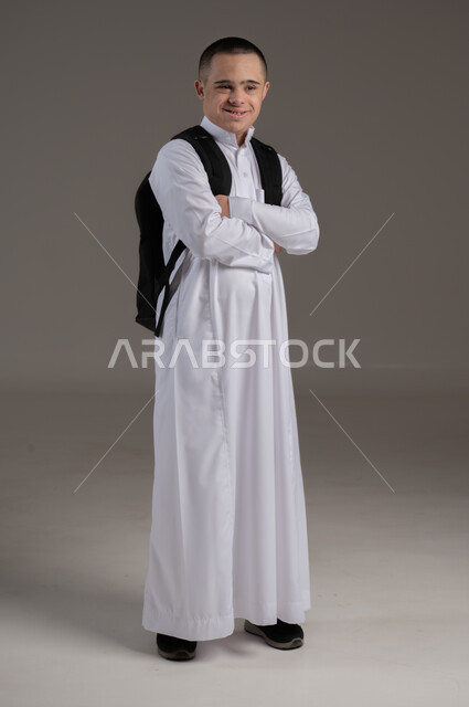مفهوم العودة الى العام الدراسي, ذوي الاحتياجات الخاصة والهمم, بورتريه لشاب عربي خليجي سعودي يرتدي الثوب وحقيبة المدرسة, الوقوف بايدي متقاطعة, صورة الجسم بالطول الكامل, خلفية رمادي