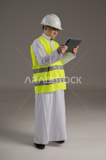 Engineering and construction works, people with special needs and determination, portrait of a Saudi Gulf Arab young man wearing a jacket and wearing a protective helmet holding a tablet in his hands, full-length body image, gray background