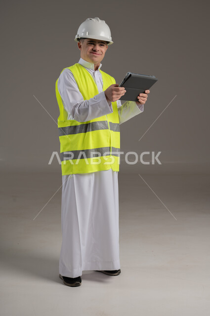 الاعمال الهندسية والبناء, ذوي الاحتياجات الخاصة والهمم, بورتريه لشاب عربي خليجي سعودي يرتدي السترة ويضع خوذة الحماية يحمل بيديه جهاز تابليت صورة الجسم بالطول الكامل, خلفية رمادي