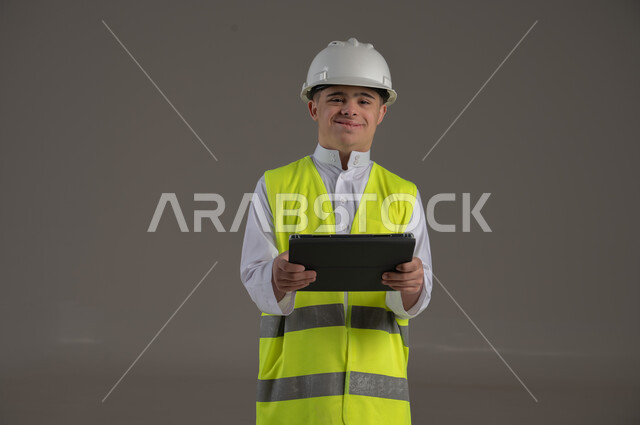 الاعمال الهندسية والبناء, ذوي الاحتياجات الخاصة والهمم, بورتريه لشاب عربي خليجي سعودي يرتدي السترة ويضع خوذة الحماية يحمل بيديه جهاز تابليت, خلفية رمادي
