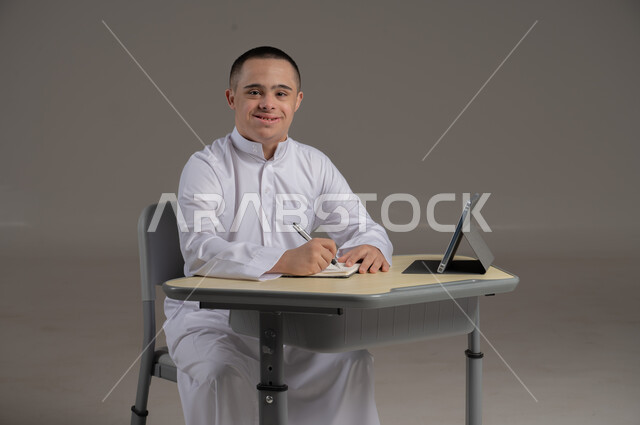 مفهوم حل الواجبات المدرسية والتعلم بواسطة التقنيات الحديثة, ذوي الاحتياجات الخاصة والهمم, بورتريه لشاب عربي خليجي سعودي يرتدي الثوب ويجلس على كرسي متحرك الدراسة بوسائل حديثة, خلفية رمادي