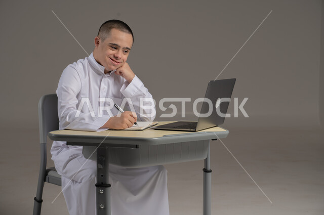 مفهوم حل الواجبات المدرسية والتعلم بواسطة التقنيات الحديثة, ذوي الاحتياجات الخاصة والهمم, بورتريه لشاب عربي خليجي سعودي يرتدي الثوب ويجلس على كرسي متحرك الدراسة بوسائل حديثة, خلفية رمادي