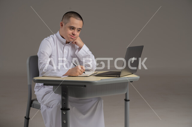مفهوم حل الواجبات المدرسية والتعلم بواسطة التقنيات الحديثة, ذوي الاحتياجات الخاصة والهمم, بورتريه لشاب عربي خليجي سعودي يرتدي الثوب ويجلس على كرسي متحرك الدراسة بوسائل حديثة, خلفية رمادي